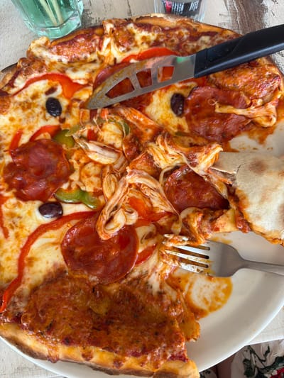 A close-up, overhead shot showcases a partially eaten pizza on a white plate. The pizza is topped with pepperoni, red and green bell peppers, olives, and shredded chicken. A metal pizza cutter and a fork are present on the plate. The background features a glass of green liquid and a light-colored wooden surface. The scene is set on a table at a restaurant in Saint-Raphaël, France.