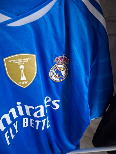 The image shows a blue Real Madrid soccer jersey laid out on a surface. The jersey has the team's crest with a crown above it, a FIFA Intercontinental Champions 2024 badge, and the Emirates "Fly Better" logo. There is no indication of the location being Batalino, Russia. The image is a close-up of the jersey's details.