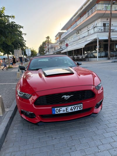 A red Ford Mustang is parked on a sidewalk next to a street with cobblestone paving. The car is facing the viewer's right and its license plate is visible, reading "OF E 4978" with a European Union emblem. The car has a black grille with a silver Mustang logo. In the background, there is a building with shops and restaurants, and trees. The sky is bright, suggesting it is daytime, likely late afternoon or early evening given the warm lighting. There are people walking on the sidewalk in the background, some appearing to be tourists. The scene appears to be in a Mediterranean coastal town. A red and white circular "no entry" sign is visible on the building in the background. The overall impression is a snapshot of a car in a pleasant, public setting during a bright day.