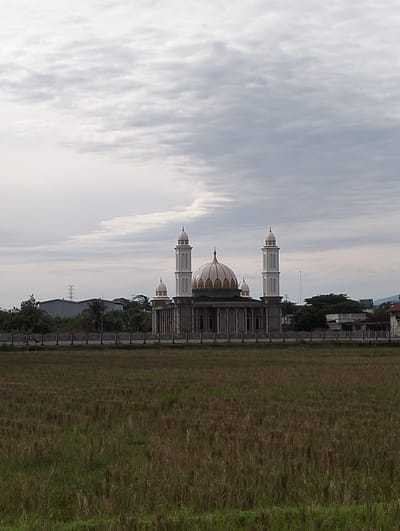 In Keudetambue, Indonesia, a mosque is depicted against a cloudy sky. The mosque features a central dome with ornate patterns and two tall minarets. The building is constructed with grey stone and has arched windows. In front of the mosque, a fence encloses the property, and beyond that, a field of dry, cut grass stretches towards the viewer. In the background, industrial buildings and trees are visible, with a power transmission tower standing out. The overall atmosphere is overcast, suggesting a subdued or possibly imminent change in weather.
