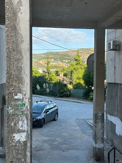 The image shows a view through a concrete structure in Балаклавский округ, Ukraine. In the foreground, two large concrete pillars frame the scene. The left pillar is heavily textured and weathered, with peeling paint and torn posters. The right pillar is smoother and also shows signs of age. Between the pillars, a paved street is visible, with a dark-colored station wagon parked on the right side of the street. The car has a roof rack. Beyond the street, there is a lush green area with trees and bushes, including some flowering plants with white blooms. In the background, rolling hills are covered with vegetation and scattered houses with red roofs. The sky is blue with scattered white clouds, and power lines are visible crisscrossing the sky. An air conditioning unit is mounted on the exterior wall of the building on the right side of the frame. The scene appears to be an outdoor urban or semi-urban environment.