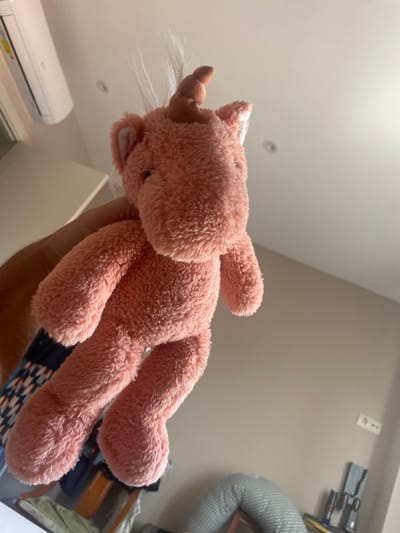 A plush toy, shaped like a pink unicorn, is held in a person's hand. The unicorn has a white horn with a white tuft of fur around it, and a white patch on its forehead. Its eyes are not visible in the image. The toy's body is fluffy and pink, with four legs. The background is a lightly colored wall and ceiling, with a light fixture visible on the ceiling. A white air conditioning unit is mounted on the wall below. In the upper right corner of the image, a gray, cylindrical object, possibly a pillow or cushion, and some electronic equipment are visible on the wall. The lighting suggests it is daytime, and the indoor setting indicates it is a domestic or indoor space, possibly in Sagunto, Spain.