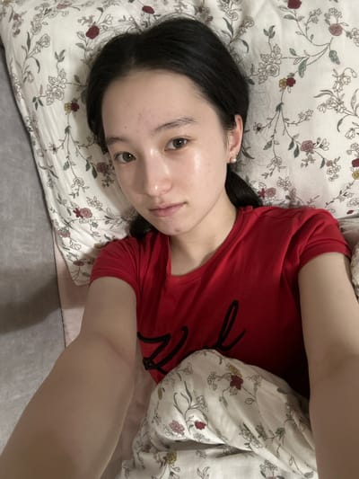 A young woman with dark hair and light brown eyes is lying in bed. She is wearing a red t-shirt and patterned pajama bottoms. The background consists of a floral pillow and a gray blanket. The scene is indoors. The location is not specified to be in the city of Nookat, Kyrgyzstan.