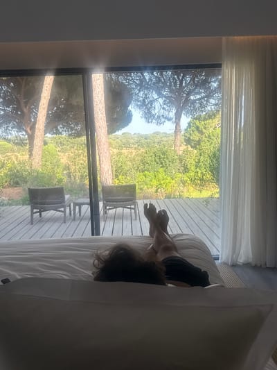 A person lies on a bed in a bedroom, facing away from the camera, looking out a large glass window at a landscape. The person's legs and feet are visible, resting on the bed. Outside the window, a wooden deck has two chairs and a small table. Beyond the deck is a natural landscape with trees and vegetation. The room has white curtains on the right side. The image is taken from a high angle, looking down at the person and the bed. The scene is bright, with sunlight coming through the window. The location is a resort in Grândola, Portugal, specifically a room with a view of the surrounding nature.
