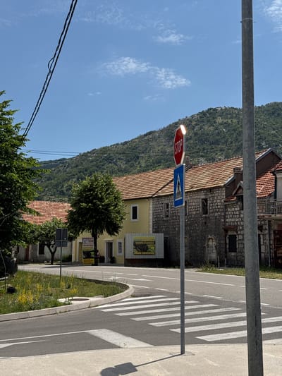 The image depicts a street scene in Erakovići, Montenegro, on a bright, sunny day. A prominent stop sign and a pedestrian crossing sign are mounted on a pole on the right side of the frame, adjacent to a crosswalk. The crosswalk stripes are clearly visible on the asphalt road, casting distinct shadows in the strong sunlight.

In the background, traditional stone buildings with tiled roofs are visible, nestled against a steep, verdant mountainside. The architecture suggests an older, European style. A yellow building with multiple windows stands out near the street. Above the buildings, a clear blue sky with scattered white clouds completes the scene.

Several objects are present: electrical wires stretch across the upper part of the frame, and trees with lush green foliage are scattered around the buildings and along the roadside. A sign with text is partially visible on the yellow building, and another sign with the words "PRŠUT-SIR" and "HOME MADE PRODUCT" is attached to a pole near the street, indicating local produce is being sold. The overall impression is a peaceful, everyday scene in a scenic, mountainous region.
