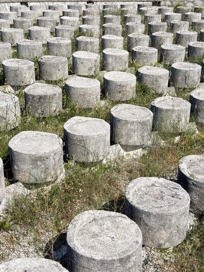 The image shows a grid of stacked, cylindrical stone blocks in Liješnje, Montenegro. The blocks are weathered and gray, with visible texture. Sparse grass and vegetation grow between the blocks. The perspective is angled, showing the receding rows of blocks. The scene appears to be a historical site or ruin. No people are visible.