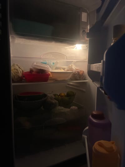 A dimly lit refrigerator is filled with various food items and containers. On the top shelf, a plastic container with food sits on top of a red bowl containing more food. To the left, a plastic bag holds produce. On the shelf below, a large bowl contains what appears to be a watermelon, and next to it, a green bowl holds citrus fruits. In the foreground, two large bottles, one purple and one orange, are visible on the door. The refrigerator is located in the city of Anggaswangi, Indonesia.