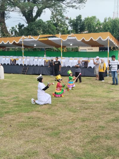 The image captures an outdoor scene in Sambirejo, Indonesia, featuring a grassy field where three children are engaged in a performance.

One child, kneeling in a white uniform and a black headscarf with a white cap, holds small batons. Two other children, dressed in bright traditional costumes with conical hats, are also kneeling and manipulating batons.

Behind the children, several adults are watching the performance, some capturing the moment with their mobile phones. A covered stage area is visible in the background, with rows of white chairs and a ladder. Trees and a telecommunications tower are also in the background.