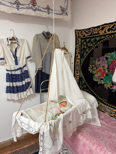 The image depicts a room, possibly in Ust'-Kabyrza, Russia, designed to resemble a historical nursery. In the center, a white, fabric-lined bassinet is suspended by ropes, containing a doll dressed in a light-colored outfit with floral patterns. The bassinet is adorned with white lace trim and embroidered details. Surrounding the bassinet, several pieces of clothing are displayed on hangers against a white wall. These include a white dress with blue trim and a plaid pattern, a grey and blue checkered dress, and another white dress with small floral embroidery. A large, dark tapestry with vibrant floral motifs hangs on the right wall. A pink patterned fabric covers a surface to the right of the bassinet, suggesting a bed or a similar piece of furniture. The overall impression is of a staged exhibit, showcasing traditional clothing and a nursery setup.