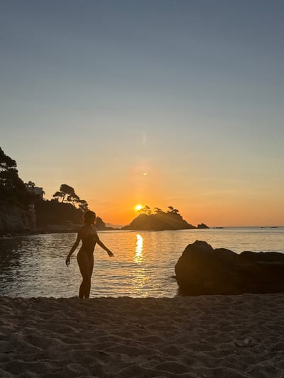 A silhouette of a woman in a dark swimsuit stands in shallow water at the edge of a sandy beach. She is facing towards a sunset that paints the sky in warm hues of orange and yellow. Her arms are extended slightly to her sides. In the background, a rocky coastline with scattered trees and buildings is visible, partially obscured by the soft light of the setting sun. The ocean reflects the sunset's glow, creating a shimmering path of light. The foreground is dominated by the textured sand of the beach. The image captures a serene moment at what appears to be Platja de Sa Conca or Platja Gran, within the municipality of Castell d'Aro, Platja d'Aro i s'Agaró, Spain.