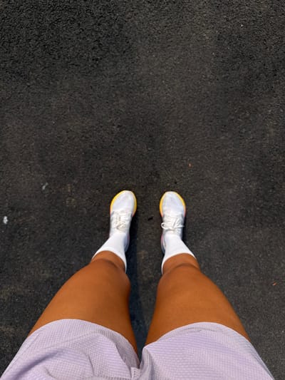 A high-angle, top-down shot captures a person's legs and feet from their own perspective, standing on a dark, textured asphalt-like surface in Marra, India. The individual wears white athletic socks and white athletic shoes, which feature vibrant yellow, pink, and blue accents along the soles. Light purple or lilac mesh athletic shorts are visible, partially covering the upper thighs. The skin tone of the legs is brown. The lighting suggests it is daytime with clear weather, suitable for outdoor activity. The scene conveys a moment of pause or preparation for physical activity, such as running or walking, given the athletic attire and surface commonly found on tracks or paths. No visible text is present on the clothing or footwear.