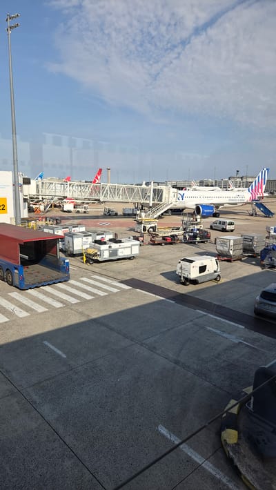 An aerial view of an airport tarmac in Tremblay-en-France, France, shows several parked airplanes and various airport vehicles. In the foreground, a blue and red baggage tug is parked. Several white baggage carts are also visible. In the mid-ground, a white passenger jet with blue and pink accents on its tail is parked at a gate, with a jet bridge extended to it. The words "Greece is bliss" are visible on the side of the plane. Another airplane with a red tail is parked in the background. The tarmac is marked with white lines and a pedestrian crossing. The sky is blue with scattered white clouds. The scene appears to be a sunny day.