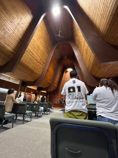 The image depicts the interior of a church, possibly located in Columbus, Ohio, United States. The architecture features a high, curved wooden ceiling with exposed beams. Rows of chairs are set up for seating, and several individuals are present, either sitting or standing. At least two people are wearing white t-shirts with writing or designs on the back. One shirt has "91" and "New York CHAMPIONS" written on it. A screen or monitor is visible in the background, displaying text in Spanish. The scene appears to be a church service or event.