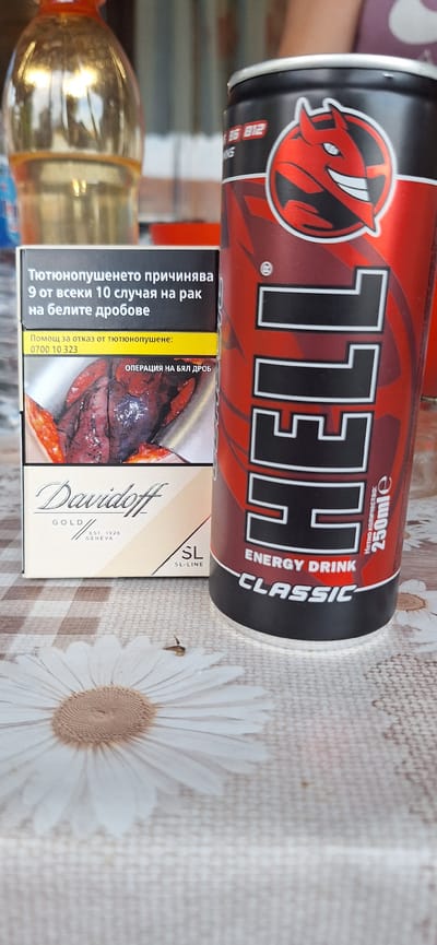 This media file captures a close-up, slightly angled view of a table setting. The primary subjects are two objects: a red and black can of "HELL ENERGY DRINK CLASSIC" and a pack of "Davidoff GOLD SL-LINE" cigarettes. The energy drink can is positioned prominently in the foreground, displaying its distinctive devil-like logo and the brand name "HELL" in large, white lettering. Text on the can indicates it contains 250ml and is an "ENERGY DRINK CLASSIC."
Adjacent to the energy drink is the cigarette pack, which features a graphic health warning depicting a diseased lung with the text "ОПЕРАЦИЯ НА БЯЛ ДРОБ" (Operation on a white lung). Other text on the pack is in Bulgarian and relates to the dangers of smoking and offers help to quit.
The background is a light-colored, textured tablecloth with a repeating pattern of white daisies. The overall lighting suggests an indoor setting, likely during the daytime, with soft light illuminating the objects. A partially visible bottle of a golden liquid, possibly a beverage, is also present to the left. There are no people visible in the frame, and the focus is entirely on the products. The scene appears to be a still life arrangement, with no specific activity or event taking place. The overall mood is neutral, with the emphasis on the commercial products and their associated health warnings.