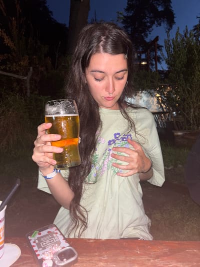 A young woman with dark hair and eyebrow piercings lies on her side outdoors. She holds a pint glass of beer with an orange slice. A light green T-shirt with purple floral pattern covers her torso. Part of a wooden table and a phone are visible on the right. The background includes trees and a hint of twilight sky.