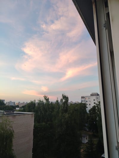 This image captures a scenic view from a window, likely in Salihorsk, Belarus, during sunset. The sky is a dominant feature, painted with soft hues of pink and pale blue, indicative of the fading light. Wispy clouds are scattered across the sky, catching the warm tones of the setting sun.
Below the sky, a dense canopy of dark green trees stretches across the mid-ground. These trees appear to be mature and full, forming a lush forest-like landscape. Amongst the trees, several apartment buildings are visible, their facades a mix of light gray and white, with numerous windows. The architecture suggests a residential area, common in Eastern European cities.
In the foreground, on the left side of the image, is a portion of a brick building, its textured surface clearly visible. On the right side, the white frame of a window occupies a significant part of the frame, framing the view and adding a sense of being indoors looking out. A small, dilapidated white structure is partially visible in the lower right, amidst the trees.
The overall atmosphere conveyed by the image is one of tranquility and the beauty of nature juxtaposed with urban living. The soft lighting and the gentle colors of the sky evoke a peaceful evening. No people or specific activities are discernible, making the scene a still-life study of a residential area at dusk. The time of day is clearly sunset or twilight. There is no visible text in the image.