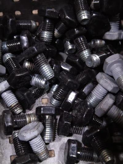 The image displays a close-up, overhead view of a large quantity of assorted bolts, predominantly black with some silver-colored ones, piled inside a dark, textured container. The bolts vary in size and thread type, with visible hexagonal heads and threaded shafts. Some bolts are partially obscured by others, creating a dense composition. The lighting is somewhat dim, casting shadows and highlighting the metallic sheen of the fasteners. There are no people, animals, or any discernible city or environmental context, including the specified location of Pargolovo, Russia, visible in the image.