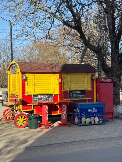 The image presents a vibrant, wagon-style food kiosk situated on a paved area in Ciorescu, Moldova. The kiosk is prominently painted in red with yellow paneling, featuring a reddish-brown shingled roof. Two black chalkboards on the front display menus in Romanian; one lists "GRILL Menu" items such as "CHEESEBURGER," "BURGER," and "CARTOFI FRI" (French fries), while the other details "BAR MENIU" options including "BERE" (beer) and "BALTURI RACOARITOARE" (refreshing drinks). An exterior lantern is mounted on the kiosk facade. A red, vertical Coca-Cola branded refrigeration unit is attached to the kiosk's right side. In front of the kiosk, on the light grey cobblestone pavement, are two red cylindrical tables with Coca-Cola branding. A dark green trash can is positioned next to these tables. To their right, a blue "Betty Ice" freezer displays various ice cream cone images. Bare deciduous trees with extensive branching occupy the background and upper portion of the frame, under a clear blue sky. A segment of a darker asphalt road is visible in the immediate foreground. Shadows indicate direct sunlight. No individuals are present in the scene.