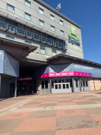 The image shows the exterior of the Hollywood Bowl and Holiday Inn Express located in Bradford, UK, at the coordinates provided. The image was taken on a sunny day, with the ground in the foreground appearing to be a brick patio. The building is light gray with numerous windows, some with balconies. The building's name, "Holiday Inn Express" is shown in blue and green letters on a sign on the exterior wall. The building is connected to Hollywood Bowl, indicated by a pink sign with the name "Hollywood Bowl". The bowl sign is displayed above the entrance to the bowling alley. The Hollywood Bowl entrance is to the left of the image, while the Holiday Inn Express is to the right. The two buildings are connected by a covered, open-air walkway, with multiple windows on the exterior of the buildings.