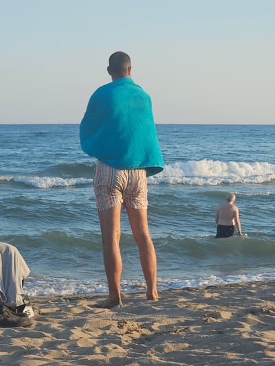 This media file captures a scene at the beach in Castelldefels, Spain. The primary focus is on two individuals. In the foreground, a person is partially visible from the waist down, wrapped in a bright turquoise towel and wearing striped swim shorts. Their legs are extended, and their feet are not clearly visible. In the background, another individual, appearing older with a lighter complexion and short hair, stands in the shallow water. This person is shirtless and wearing dark swim shorts.

The activity depicted is a day at the beach, with one person presumably drying off or resting near the shore and the other wading in the ocean. The setting is a sandy beach with the Mediterranean Sea in the background. Waves are breaking near the shore, indicating moderate sea conditions. The sand is textured with footprints and ripples, suggesting it's a well-used area. The lighting implies it is daytime, likely mid-morning or late afternoon, given the angle of the sun casting some shadows on the sand. The overall mood appears relaxed and recreational.

There are no discernible emotions conveyed through the visible parts of the subjects. Notable details include the distinct color of the towel and the pattern of the swim shorts. A small, black object with some white markings is visible on the sand in the lower right corner of the frame, possibly a bag or container, but the markings are too indistinct to read any text. The clear blue sky and the vibrant blue of the sea suggest fair weather conditions, suitable for a beach outing.