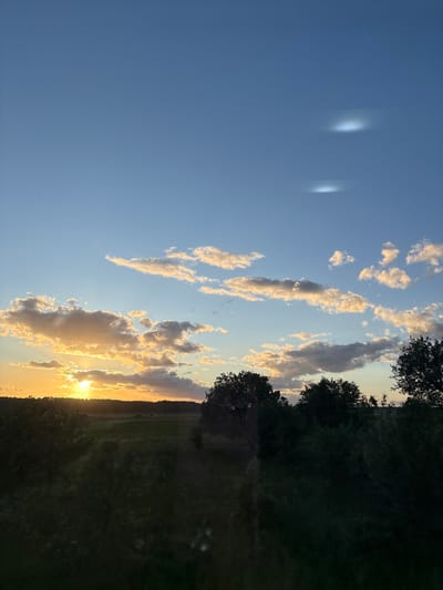 The image depicts a landscape at sunset, possibly in Мажэйкаўскі сельскі Савет, Belarus. The sky, which takes up a significant portion of the frame, is a clear blue with scattered clouds tinged orange by the setting sun. The sun itself is low on the horizon, partially obscured by clouds, creating a bright golden glow. The lower portion of the image shows a dark, grassy area, possibly a field, with a line of trees visible along the horizon. The image is oriented vertically. There are a few light artifacts on the left side of the image.