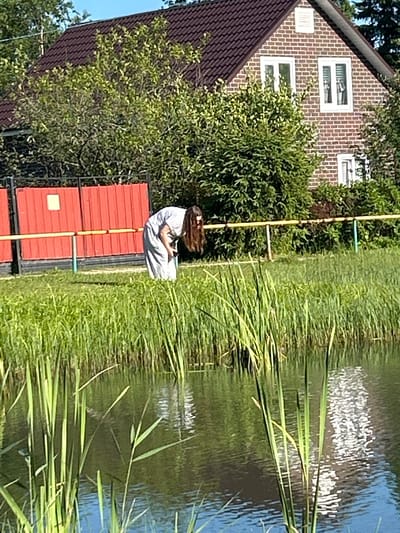 A woman in a light blue dress is bending over near the edge of a body of water. She has long, dark hair that falls forward, obscuring her face. The water has tall reeds growing along its edge, and a reflection of the sky and greenery is visible on the surface. Behind the woman, there is a red fence with yellow and grey poles. Further back, a brick house with a dark tiled roof and white-framed windows is visible, surrounded by lush green trees and bushes. The scene appears to be in a suburban or rural setting.