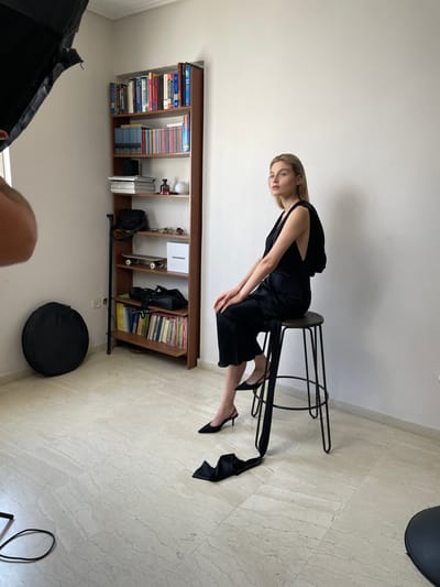 The original image is rotated 90 degrees clockwise. When viewed correctly, it depicts an indoor, full-shot photograph of a young woman posing for a photoshoot, likely in a studio or a well-lit room in Vironas, Greece. The woman is positioned slightly off-center to the right, seated on a tall, black metal stool with a dark wooden top.
The woman has long, straight blonde hair and is dressed in a sleek black sleeveless garment that features a draped or flowing fabric detail from the back, extending towards the floor. She wears black pointed-toe high-heeled mules. She leans forward on the stool, resting her right hand on her left knee, and looks directly towards the camera with a neutral, composed expression. Her left leg is subtly extended, with the black fabric detail from her garment trailing near her foot.
The background features plain, light-colored walls and a light-tiled floor with a subtle pattern. To the left, a wooden bookshelf is visible, well-stocked with numerous books, some small decorative items, and a prominent white box labeled "PROGRESS." On the floor to the lower left, a black circular photography softbox or reflector is present, alongside part of another piece of photography equipment in the bottom-left corner, suggesting a professional photography setup. The lighting appears to be controlled studio lighting rather than natural daylight.