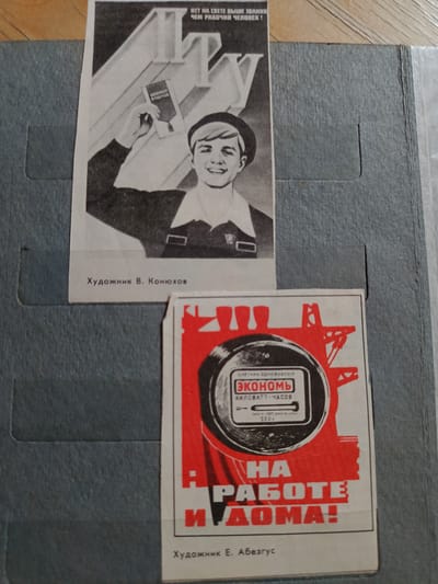 The image shows two posters within a blue binder. The top poster is a black and white illustration of a smiling young man holding up a small rectangular object. Above him are large stylized letters. Text below the illustration reads "Художник В. Конюхов".
The bottom poster is predominantly red and black, featuring a stylized drawing of an electric meter with the word "ЭКОНОМЬ" (Economize) in large letters. Below this is text that reads "НА РАБОТЕ И ДОМА!" (At work and at home!). Text at the bottom reads "Художник Е. Абезгус". The binder appears to be made of blue cardboard and is lying flat on a wooden surface. There are no recognizable landmarks or details that would indicate the location in Bugra, Russia.