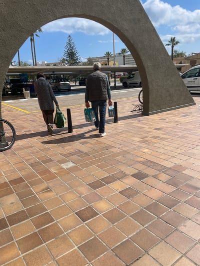 An overhead concrete archway frames a sunny outdoor scene in Sant Llorenç des Cardassar, Spain. The ground in the foreground and beneath the archway is paved with varied shades of terracotta-colored square tiles. Two individuals, observed from behind, walk away from the viewer through the archway onto a paved parking area. The person on the left wears a grey patterned long-sleeved top and dark trousers, carrying a green reusable shopping bag in their right hand. The person on the right, an adult male with grey hair, wears a dark olive vest over a blue long-sleeved shirt and blue jeans, holding a multi-pack of bottled beverages in their left hand. Both individuals cast distinct shadows to their right, indicating a light source from the left.
Beyond the archway, a spacious asphalt parking lot is visible, partially covered by white overhead shade structures. Multiple vehicles are parked, including a silver hatchback, a white commercial van with "coemsa" branding, and other cars. A designated area with shopping carts from "Carrefour" is visible to the left. Several black cylindrical bollards line the edge of the tiled path separating it from the parking area. A partial view of a bicycle wheel is visible on the far left foreground, and a bicycle rack is situated to the right. The background features various buildings, including one with a "BLUESEA" sign. Tall palm trees and a prominent conical conifer tree punctuate the landscape against a clear blue sky with scattered white clouds.