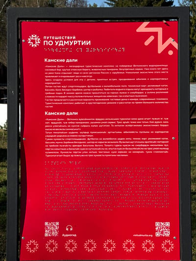 The image shows a red sign with white text in Russian, set in a black metal frame. The sign is titled "ПУТЕШЕСТВУЙ ПО УДМУРТИИ" ("TRAVEL TO UDMURTIA") at the top, followed by "Камские дали" ("Kama Dal") written in a larger font. The sign contains information about the "Kama Dal" tourist complex on the Votkinsk reservoir in Russia, describing its features, activities, and amenities. The text is also provided in braille for visually impaired individuals. The lower portion of the sign has an audio guide icon, a QR code, and the website address "visitudmurtia.org". Star-shaped designs adorn the right side of the sign. The surrounding environment appears to be outdoors, possibly in a park or recreational area, with trees visible in the background.