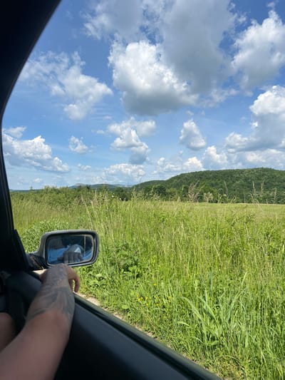 The image is a view from inside a vehicle, possibly a car, looking outwards through an open window. A portion of a grassy field occupies the right side of the frame, and in the background, a treeline rises, suggesting a hill or mountain range. The left side of the frame is dominated by a bright blue sky populated with puffy white clouds. The vehicle's side mirror is visible, reflecting a hand and a hint of the person's shirt. A tattooed arm extends into the frame. The location is in the vicinity of Kastel, Bulgaria, identifiable through visual cues suggesting a rural environment and the terrain.