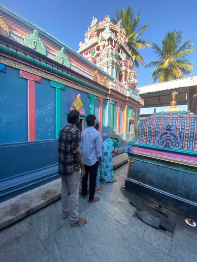In Chinnambedu, India, at a temple, three individuals stand on a stone courtyard. Two men, facing away from the camera, are dressed in casual attire. One man wears a plaid shirt and light-colored pants, while the other is in a light-colored button-down shirt and dark pants. A woman in a green and white floral dress is also present, her back to the camera. She appears to be bending or kneeling. The temple structure features vibrant, multi-colored decorations, including intricate carvings and painted details on its towers and walls. Palm trees are visible against a clear blue sky. The ground is paved with stone tiles, and a dark, rectangular structure with decorative elements is positioned to the right of the frame. A small white plate with offerings is on the ground near the dark structure. The scene suggests a religious or cultural gathering at the temple.