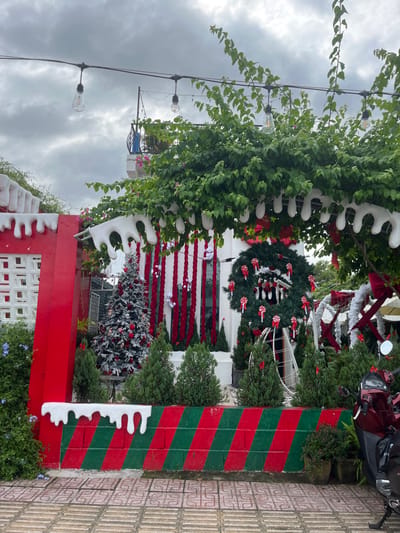A festive scene showcases a building or entrance adorned with Christmas decorations. A large wreath with candy cane accents hangs above, while vertical red and white garlands drape below. A decorated Christmas tree is also visible. Lush greenery with decorative white elements frames the structure. A pole painted with diagonal red and green stripes stands adjacent to a tiled walkway. The sky is overcast.