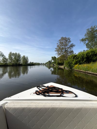 View from a boat on a canal in Rhenoy, Netherlands. The canal is bordered by trees and foliage, their reflections visible in the water. A boat cleat with rope is attached to the side of the boat, adjacent to a white quilted seat. The sky above is blue.