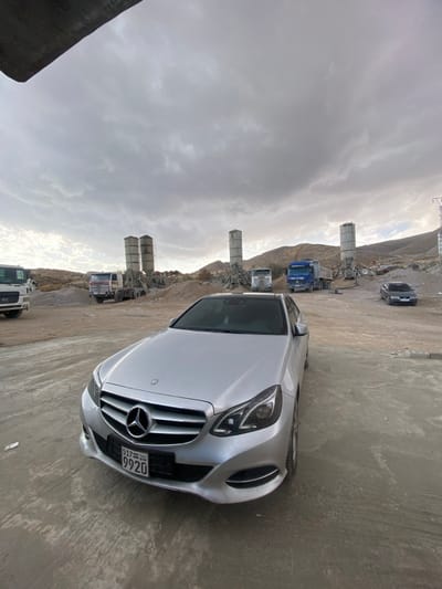 The image depicts an outdoor industrial scene located in Dahiyat Harasta, Syria, under an overcast sky. In the immediate foreground, a silver Mercedes-Benz E-Class sedan (W212 facelift) is parked facing the viewer at a slight angle to the right. The Syrian license plate, "537 SYR 9920", is clearly visible on the front bumper. The ground beneath the vehicle is uneven, consisting of packed dirt and concrete.
In the mid-ground, an active or recently active aggregate processing or concrete batching plant is visible. This includes multiple tall, cylindrical structures, likely cement silos or aggregate hoppers, arranged in two distinct pairs. Surrounding these structures are large piles of various aggregates, such as sand and gravel. Several heavy vehicles are present: a white utility truck is partially visible on the far left, another white truck (possibly a concrete mixer) is positioned behind the left pair of silos, and a blue dump truck is to the right of this pair. A second white truck is visible further right, near the second pair of silos. A dark-colored sedan is parked further back on the right side of the scene.
The background consists of arid, rocky hills or mountains under a heavily clouded, grey sky. No individuals are visible, and no actions or interactions are depicted; all vehicles appear stationary. A power line pylon or structure is partially visible on the far right.