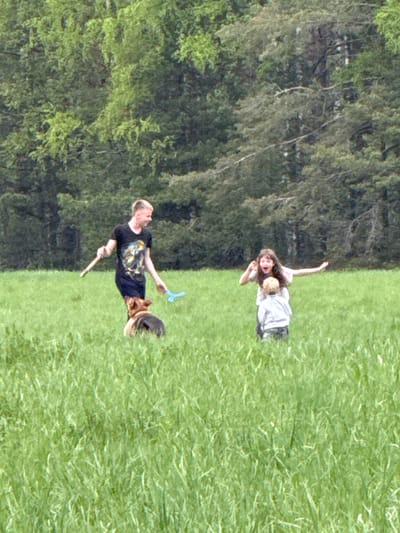 The image depicts a grassy field adjacent to a wooded area. A man wearing a black t-shirt and black shorts is holding a blue object in his right hand and a stick in his left. A woman with long dark hair and a child are standing together, with the woman's right arm raised. The location is Малая Рукавицкая, Russia.