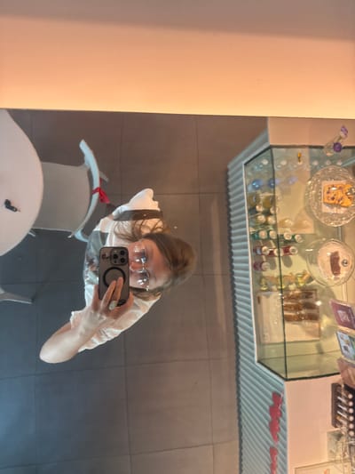 A woman takes a selfie in a mirror, reflecting a cafe display case filled with pastries and drinks. The display case is lined with a white, corrugated material. The woman is wearing glasses and a light-colored top. She holds a smartphone up to her face. Below her reflection is a white table and chair. A small portion of a wall is visible on the left side of the frame.
