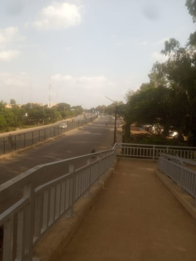 The image depicts a street scene in Unguwan Dosa, Nigeria, during the daytime. The perspective is from an elevated walkway with a concrete path and a metal railing on either side, looking down onto a road. The road appears to be a multi-lane street with a black asphalt surface. Traffic is visible on the road, including cars, motorcycles, and what appears to be a three-wheeled auto-rickshaw. Vegetation, primarily trees, lines the sides of the road, with some buildings visible in the background, suggesting an urban or semi-urban environment. Several tall communication towers are also visible in the distance. The sky is mostly clear with scattered white clouds, indicating fair weather. The lighting suggests it is likely morning or late afternoon. There is no visible text in the image.
