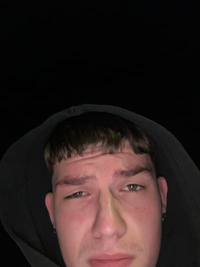 A close-up, low-angle, indoor shot shows a young person wearing a black hooded sweatshirt, their face illuminated by a light source from above and slightly to the left. The background is completely black, suggesting a dimly lit or dark environment. The person has short, dark brown hair with bangs, light brown eyes, and fair skin with some redness on the cheeks. Their eyebrows are dark and well-defined. They are wearing a small hoop earring in their visible left ear. Their facial expression appears somewhat pensive or perhaps slightly uncomfortable, with a furrowed brow and slightly downcast eyes. The image is cropped such that only the upper portion of the person's face and head are visible. The specific location in the city of Siedlce, Poland cannot be determined from the visual information provided.