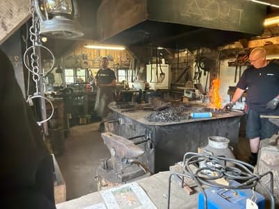 The image depicts a blacksmith's workshop in Lejre, Denmark, with two men actively engaged in their craft. In the foreground, a large anvil sits on a wooden base. Behind it, a robust workbench is piled with metal scraps and tools, with a gas torch or forge actively burning, casting a vibrant orange flame. One man, wearing a black t-shirt and shorts, stands at the workbench, holding tongs and working with the glowing metal. Another man, wearing a dark t-shirt and apron, stands to the left, observing the process. The workshop environment is rustic, with wooden beams, hanging lanterns, various tools, and a large ventilation hood above the forge. The overall atmosphere suggests a traditional and working blacksmith's studio.