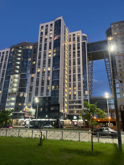 The image captures a modern apartment complex in the city of Dachnoe, Russia, at twilight. The multi-story buildings, characterized by a combination of white panels and dark blue glass facades, are illuminated by numerous yellow lights visible through the windows. A prominent architectural feature is a skybridge connecting two sections of the buildings, creating an open, arch-like structure against the darkening blue sky. Streetlights are visible, casting a bright glow, and a utility pole with wires stands to the right of the frame. Below the buildings, a street is active with car traffic, including a dark sedan moving past. A metal fence separates the street from a grassy area in the foreground, where a young tree stands. A billboard with text in Russian is mounted on the side of one of the buildings.
