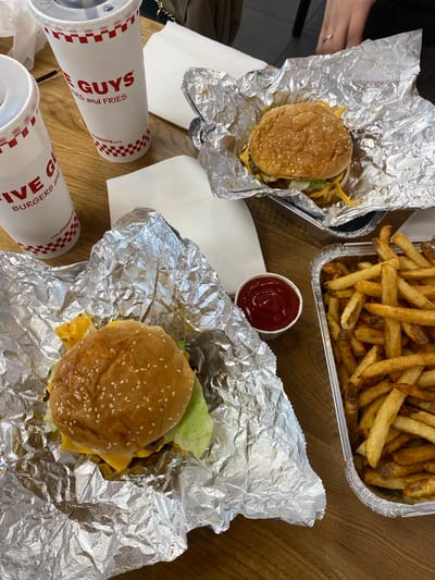 The image shows a table setting, likely in a restaurant or fast-food establishment, in Uppsala, Sweden. The food present consists of two hamburgers wrapped in foil, a container of french fries, and a small cup of ketchup. There are also two cups with lids, branded with "FIVE GUYS BURGERS and FRIES". The table surface is made of light brown wood. The presence of food wrappers and containers suggests a recent meal. The lighting appears to be natural. The image also shows a woman's hands in the upper left corner.