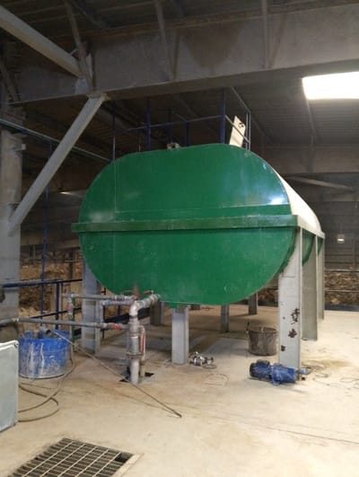 The image shows a large, green, cylindrical tank in a factory setting. The tank has a cylindrical bottom and a curved top. There are four steel support columns extending from the bottom of the tank. The tank appears to be resting in a concrete area, with a metal grate in the foreground. Several pipes and valves can be seen under the tank, connecting to the steel support columns. There is a blue bucket in the foreground, and a smaller bucket and a blue electric motor are visible in the background. The location of the scene is a factory in Pangasia, Bangladesh, based on the provided coordinates (latitude: 22.693, longitude: 90.601).  There are no people in the image.