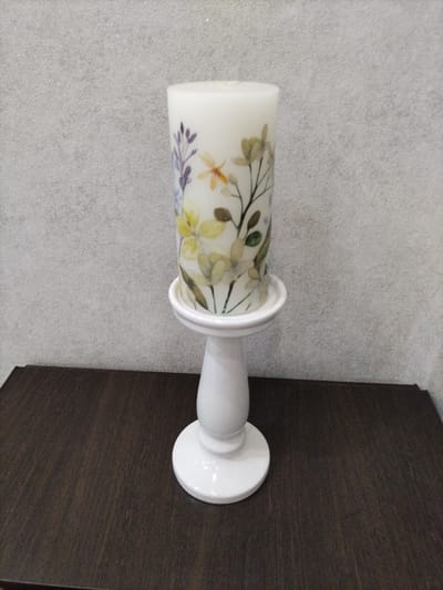 A white cylindrical candle adorned with a delicate floral and dragonfly motif stands on a white ceramic candlestick. The candlestick has a round base, a stemmed middle section, and a small dish to hold the candle. The candle and candlestick are positioned on a dark, wooden surface. In the background, a textured, light gray wall serves as the backdrop. The scene appears to be an indoor setting, possibly a home or a decorative display. The location is not specified in the image.