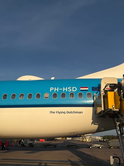 A KLM Boeing 737-800 aircraft, registered PH-HSD, is parked on the tarmac at Stockholm-Arlanda Airport, Sweden. The aircraft displays a blue upper fuselage and a white lower fuselage, separated by a thin white stripe. The registration PH-HSD is visible in white on the blue section, flanked by the Dutch flag (red, white, blue horizontal) and the European Union flag (yellow stars on blue). Below the windows, the phrase "The Flying Dutchman" is printed in black. A mobile yellow and grey passenger stairs unit is positioned at the aircraft's forward boarding door, with one individual partially visible on the top platform. On the dark asphalt tarmac, a white ground support vehicle marked "TRU 246" is pulling a string of empty baggage dollies. Another individual, dressed in dark work attire, is visible on the tarmac near the aircraft's belly. The background includes distant airport buildings, additional ground equipment, and a line of trees under a clear, bright blue sky. Natural sunlight illuminates the scene, casting distinct shadows on the ground.