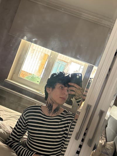 This image displays a young male taking a mirror selfie inside a room. The specific city of Tatishchevo, Russia, cannot be visually confirmed from the image content.
The subject has dark, curly hair and is wearing a long-sleeved, horizontal striped shirt (dark and light). He exhibits distinct black tribal-style tattoos on both sides of his neck and patterned tattoos, resembling branches, on his visible right forearm. He holds a dark-colored smartphone, likely an iPhone due to the visible camera array, with his left hand, partially obscuring his face. His gaze is directed slightly away from the phone's lens, towards his reflection.
He is positioned on a bed with light-colored bedding. The mirror used for the selfie features a light-colored frame and two silver horizontal handles on the right side, suggesting it is part of a piece of furniture like a wardrobe or closet door. Behind the subject, a window with a white frame is visible, partially covered by a grey roller blind. Through the window, an exterior view includes parts of a building or structure with exposed wooden elements and a segment of a light blue painted surface. A large, bright green moth, possibly a Luna Moth, is visible on or near the windowsill. The interior wall to the left of the window exhibits a textured, grey finish. The room is illuminated by natural light entering through the window.