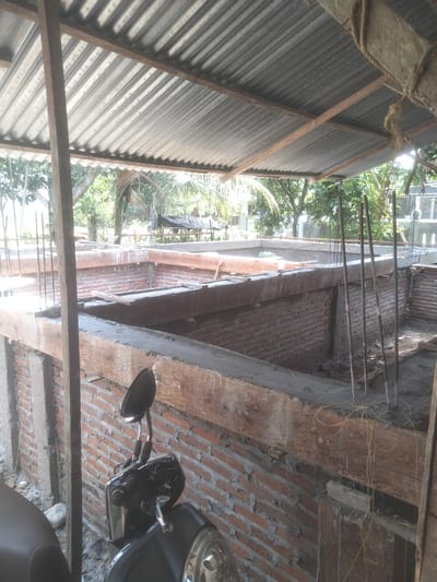 The image shows a construction site in Kruengmanyang, Indonesia. The foundation of a building is under construction, with red brick walls and concrete beams. Rebar rods protrude vertically from the concrete. The construction is covered by a corrugated metal roof supported by wooden beams. A black motorcycle is parked in the foreground. Trees are visible in the background outside the construction area.