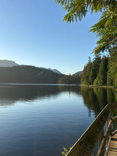 The image depicts a tranquil lake scene with a wooden walkway in the foreground. The lake is calm, reflecting the clear blue sky and the surrounding evergreen forest. In the distance, mountains are visible, partially obscured by the trees. The sun appears to be low in the sky, casting a gentle light on the landscape. This scene is located in Belcarra Regional Park, within the city of Port Moody, Canada. There are no people or animals visible in the image.