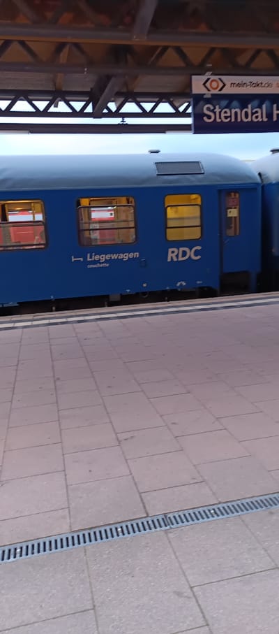 The image shows a side view of a blue train car with the words "Liegewagen" and "RDC" printed on its side. The train is stopped at a station platform paved with light-colored square tiles. Above the train, a sign reads "mein-Takt.de" and "Stendal Hbf" which indicates Stendal Hauptbahnhof (main train station), in Stendal, Germany. The train car has several windows, some of which are lit from within, suggesting the interior is illuminated. The station appears to be covered by a roof structure supported by metal beams. A metal grate runs along the edge of the platform. The overall lighting suggests it might be early morning or late evening. There are no people clearly visible in the image.