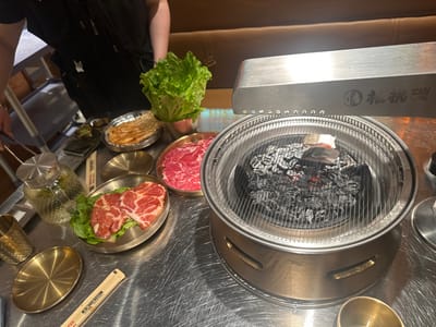 A dining table holds a Korean BBQ setup. Raw meat, lettuce, and side dishes in small brass bowls surround a round grill with hot coals. An overhead vent hood partially obscures the grill. Chopsticks and a small brass cup are on the table. A person wearing a black apron stands on the left side. The table surface is stainless steel.